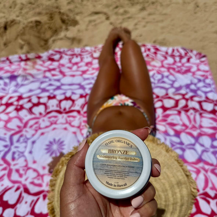 Hand holding a jar of bronzer balm on a beach with sand and towel in the background