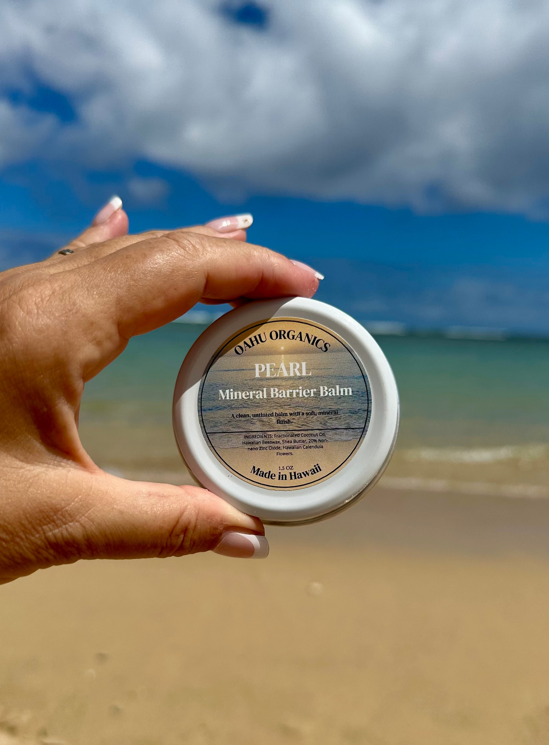Hand holding a jar of Pearl Mineral Barrier Balm on a beach with ocean and sky in the background
