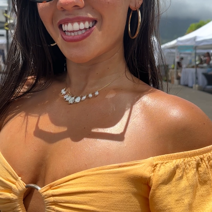 A woman outdoors wearing a mustard yellow off-the-shoulder top, black sunglasses, and gold hoop earrings, holding a bottle of Oahu organics shimmery oil.